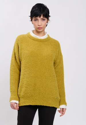 Mujer con cabello negro corto que lleva un jersey texturizado de color mostaza sobre una camisa blanca y pantalones negros, de pie frente a un fondo liso.