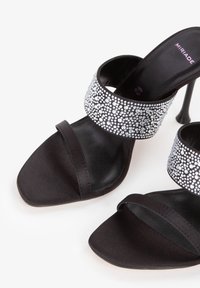 Black open-toe high-heeled sandals with two straps, one adorned with sparkling rhinestones, on a white background.