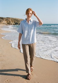Jeune femme aux cheveux roux marche pieds nus sur une plage de sable près des vagues, portant une chemise bleu clair, un pantalon beige et des sandales marron sous un ciel dégagé.