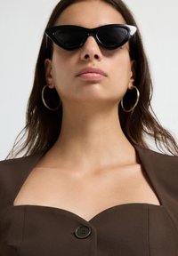 Vrouw die zwarte cat-eye zonnebril, grote gouden oorringen en een bruin off-shoulder top met een knoop in het midden draagt.