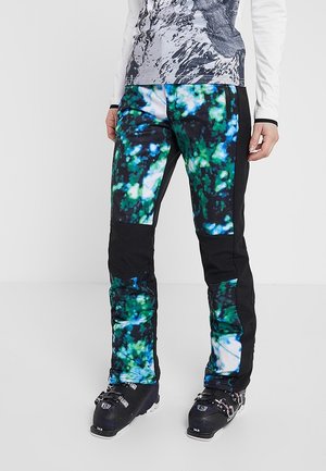 Person iført farverige skis, der er mønstret med abstrakte former, og sorte skistøvler, stående på en ensartet lys baggrund.