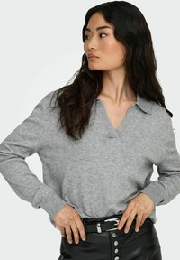 Grau, leichter Pullover mit V-Ausschnitt und langen Ärmeln, strukturierter Strick, kombiniert mit einem schwarzen Lederrock mit silbernen Knöpfen und einem Gürtel.