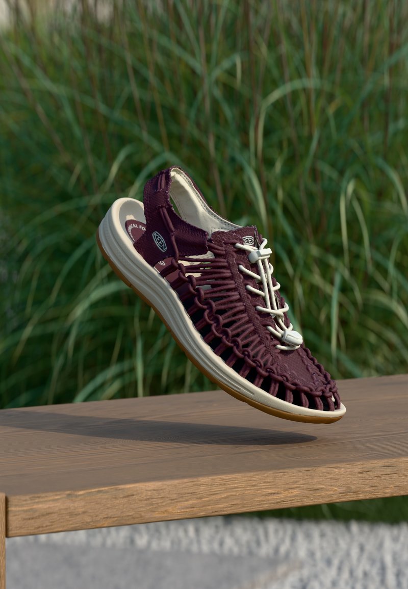 Μπορντό υφασμένο σανδάλι με λευκά κορδόνια και μπεζ σόλα, κρεμασμένο πάνω από ξύλινο παγκάκι με φόντο πράσινο γρασίδι.