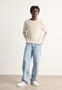 Beige knit sweater with a round neckline, paired with light blue denim jeans. White sneakers complete the outfit. Simple design, relaxed fit.