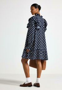 Marineblå dottedress med flæser på skuldrene, lagdelt nederdel og lange ærmer. Båret med en brun taske og brune sko, der har hvide sokker.