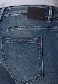 Jeans de mezclilla azul oscuro con un acabado texturizado, que cuentan con una etiqueta de cuero negro y un bolsillo trasero con costuras visibles.