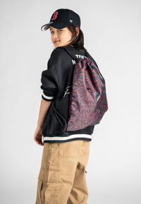 Mladý člověk na sobě má černou baseballovou kšiltovku, černou mikinu, béžové cargo kalhoty a černo-červený batoh se geometrickým vzorem na stažení.