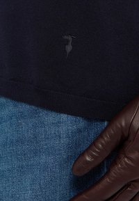 Maglione a maglia blu navy con un piccolo logo ricamato scuro, abbinato a jeans blu e un guanto in pelle marrone che afferra il tessuto.