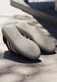 Scarpe da ginnastica nere con suola bianca e strisce bianche, posate con la suola rivolta verso l’alto su una superficie di cemento illuminata dal sole con motivi di ombre.