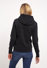 Sudadera negra con una gran capucha, puños y cintura acanalados, que presenta una textura suave y un diseño casual. Llevada con jeans azul.