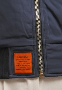 Marine bomberjack met een gouden rits, zwarte geribde manchetten en een oranje label dat de specificaties van de U.S. Air Force vermeldt.