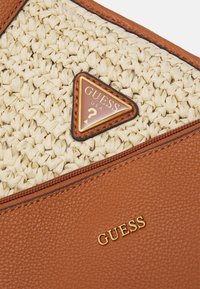 Section de sac en tissu beige tissé et cuir marron texturé, avec le logo Guess en lettres dorées et un emblème triangulaire Guess USA.