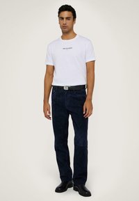 Camiseta de algodón blanca con impresión de "TRUSSARDI", combinada con pantalones de ante azul marino y botas de cuero negras. Diseño simple y ajustado.