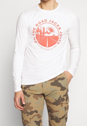 Chemise blanche à manches longues avec un motif graphique rouge représentant un paysage désertique et du texte. Associée à un pantalon cargo camouflage kaki.