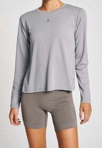 Langärmliges graues Sportshirt mit Rundhalsausschnitt, das ein kleines Logo und eine glatte Textur aufweist, kombiniert mit hellbraunen, eng anliegenden Shorts.