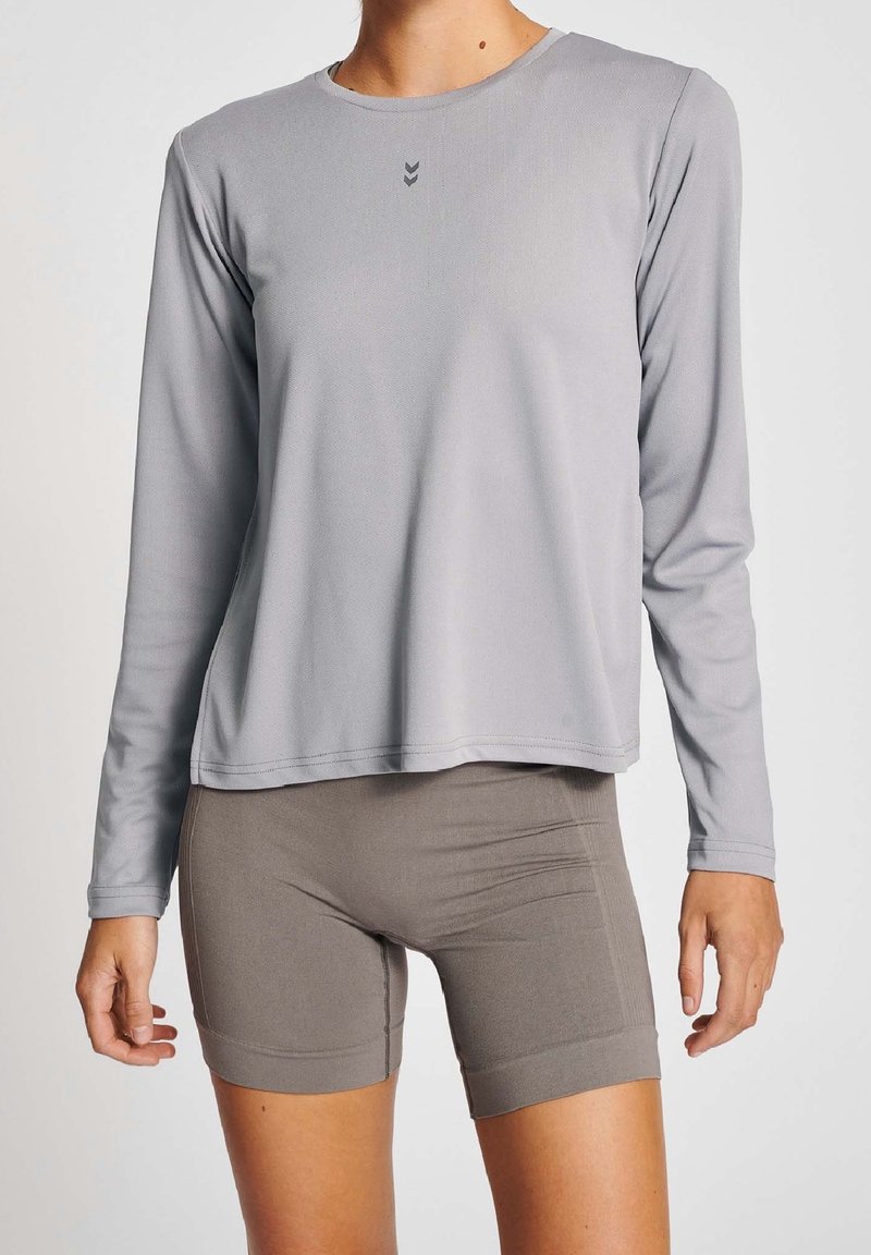 Langärmliges graues Sportshirt mit Rundhalsausschnitt, das ein kleines Logo und eine glatte Textur aufweist, kombiniert mit hellbraunen, eng anliegenden Shorts.