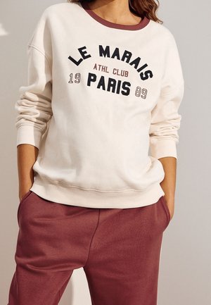 Kvinde iført en cremefarvet sweatshirt med teksten "LE MARAIS ATHL CLUB PARIS 1989" og vinrøde joggingbukser med hænderne i lommerne.
