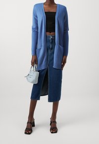 Mujer con cardigan largo azul, top negro sin mangas, falda midi de mezclilla con abertura frontal, sandalias negras de tacón, sosteniendo un bolso pequeño azul claro.