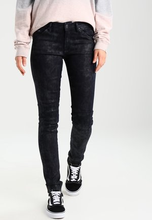 Osoba v černých přiléhavých texturovaných kalhotách, černo-bílých teniskách a světle růžovo-šedém přechodovém dlouhém rukávu, kráčející vpřed.