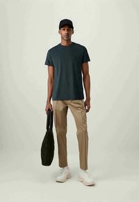 Dark green short-sleeve t-shirt, beige slim-fit trousers, white sneakers, and a black textured tote bag. Model wears a black baseball cap.