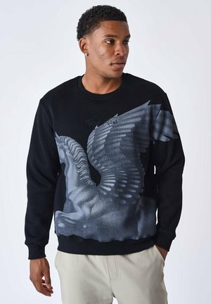 Sweatshirt noir avec un grand motif de cheval ailé gris, col rond et poignets côtelés, assorti à un pantalon de couleur claire.