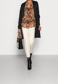 Manteau noir par-dessus un blouse fleurie marron, pantalon blanc et bottines noires à talons. Tenant un sac à main noir avec des poignées beige.