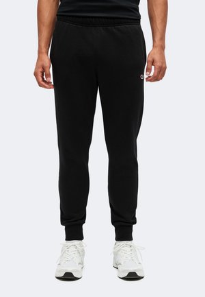 Un modèle porte un pantalon de jogging noir avec des bas resserrés aux chevilles et un petit logo, associé à des baskets blanches.