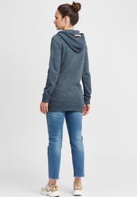 Grå hettegenser med snøring på hetten, lange ermer og ribbestrikkedemansjetter. Sammen med lyseblå jeans og beige sneakers. Glatt tekstur.