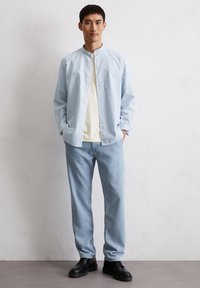 Camicia a righe azzurro chiaro con colletto alla mandarina, abbinata a una maglietta crema e jeans azzurro chiaro. Scarpe nere completano l'outfit.