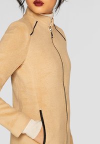 Beige, textured outerwear with a high collar and zip closure, featuring dark accent seams and side pockets. Soft wool-like material.