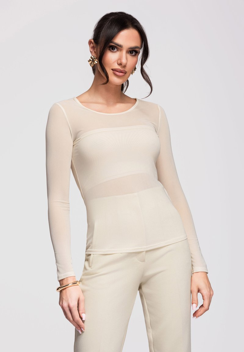 Femme aux cheveux noirs portant un haut beige transparent à manches longues et un pantalon assorti, avec des boucles d'oreilles et bracelets en or, posant devant un fond uni.
