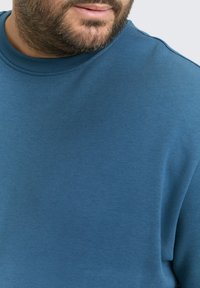 Niebieska bluza z gładkiego materiału, z okrągłym dekoltem i długimi rękawami. Zbliżenie ukazuje luźny krój z subtelnie zaznaczonymi szwami.