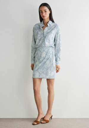 Donna che indossa un vestito camicia a righe azzurro chiaro con motivi floreali bianchi e sandali marroni aperti con tacco, in piedi contro un muro semplice.