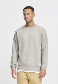Ung mann iført lys grå sweatshirt og beige bukser, stående mot en enkel hvit bakgrunn, ser fremover med nøytral uttrykk.