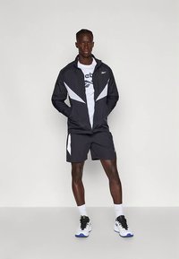 Άνδρας που φοράει αθλητική ζακέτα σε μαύρο και λευκό χρώμα, αντίστοιχα σορτς, λευκό μπλουζάκι, λευκές κάλτσες και λευκά αθλητικά παπούτσια με μπλε σόλες, στέκεται με τα χέρια στις τσέπες.