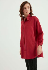 Rotes Hemd mit langen Ärmeln, aus weichem Stoff, mit einem Kragen, großen vorderen Knöpfen und einem abgerundeten Saum. Getragen über schwarzen Hosen.