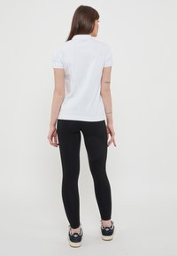 Polo bianco a maniche corte, con design aderente, indossato con leggings neri. Il modella ha i capelli lunghi e sta girata di spalle rispetto alla telecamera.