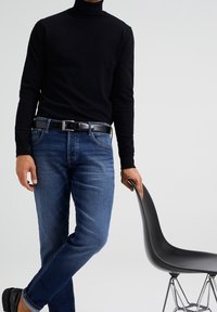 Svart polotröja, mörkblå jeans, svart bälte och svarta skor. Personen lutar sig mot en modern svart stol med en böjd sits.