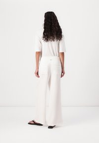 Pantaloni bianchi, larghi e svasati, con un design a gamba larga, caratterizzati da una vita arricciata e due tasche sul retro. Indossati con ciabatte nere.