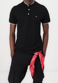 Zwarte poloshirt van katoen met korte mouwen, witte kraagafwerking, knoopsluiting met twee knopen en klein logo; rode bandana geknoopt om de taille.