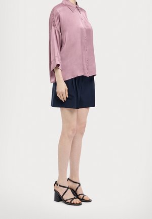 Staande persoon die een loszittend roze overhemd met knopen, donkerblauwe korte broek en zwarte sandalen met hak en enkelbandjes draagt.