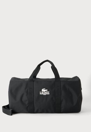 Sort Lacoste rejsetaske med to håndtag og en aftagelig skulderrem, med Lacoste-logoet i midten foran.