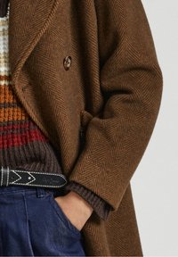 Cappotto marrone a spina di pesce con design doppiopetto, indossato sopra un maglione a righe in maglia. Include una cintura nera con cuciture bianche.