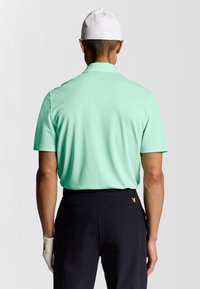 Lichtgroene polo met een zachte textuur, korte mouwen en een geribde kraag, gecombineerd met donkere broeken met een geel logo op de achterkant.