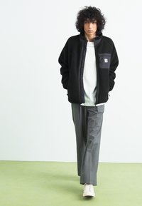 Sort fleecejakke med lynlås foran og sidelommer, parret med grå bukser og lyse sneakers på et grønt gulv.
