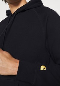 Persona che indossa un cappuccio nero con stringhe, mostrando parzialmente una mano e un piccolo logo giallo sul polsino della manica.