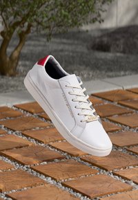 Sneaker bianca con una superficie liscia, dettagli rossi sul tallone, lacci beige e un logo dorato sul lato, posata su piastrelle di legno.