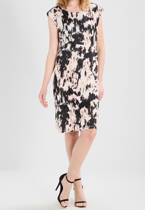 Vrouw draagt een knielange mouwloze jurk met abstracte zwarte en beige print, gecombineerd met zwarte hoge hakken met enkelriem.