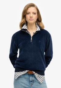 Sudadera de forro polar azul marino con cremallera de cuarto, con dobladillo y puños acanalados. Se lleva sobre una camisa de collar a rayas. Textura suave.