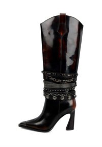 Bota de cuero negro hasta la rodilla con punta afilada, que presenta correas metálicas con tachuelas y cadenas alrededor del tobillo. Diseño de tacón de aguja.
